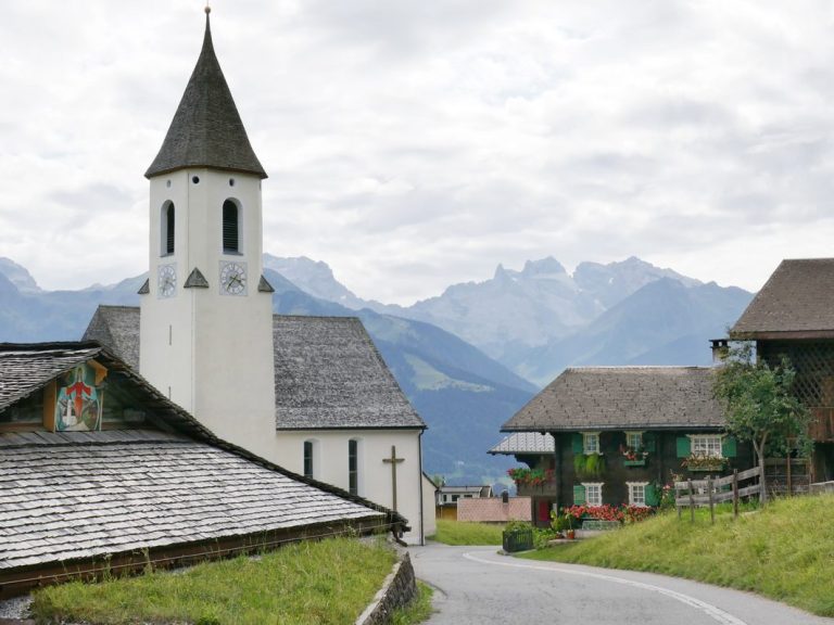 Bellavista-Brand_Freizeit-Ausfluege Vorarlberg Landschaft mit Kirche, traditionellen Häusern und Bergkulisse unter bewölktem Himmel.